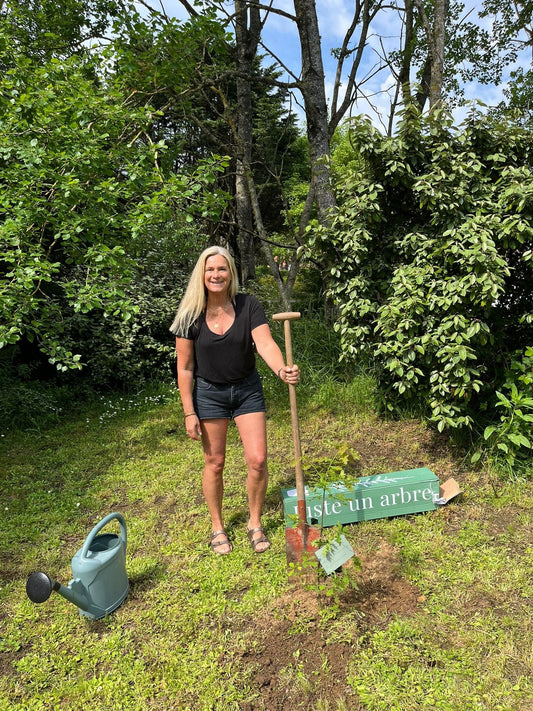 Jeune couple souriant plantant un olivier dans leur jardin pour célébrer leur emménagement, entouré d’outils de jardinage et d’une plaque gravée posée au pied de l’arbre.